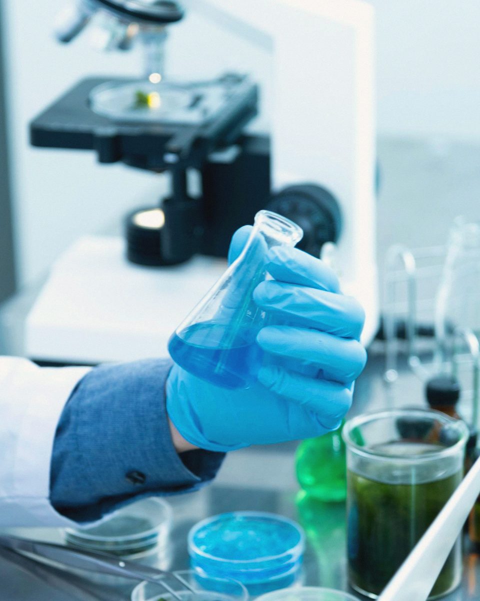 A gloved hand holding a flask of blue liquid in a laboratory, microscope behind.
